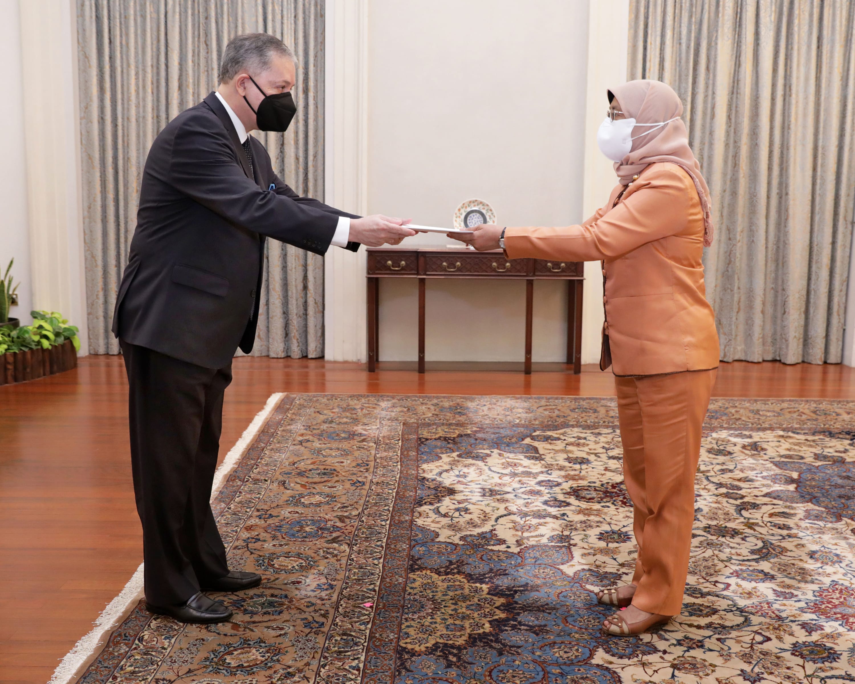 Man in suit and woman in hijab exchanging papers, both wearing face masks.
